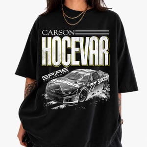 May include: A black short-sleeve t-shirt featuring a graphic print of a race car with the number 77. The text reads CARSON HOCEVAR and SPIRE MOTORSPORTS. The design includes a black and white race car with the word Spectrum on the side panels.