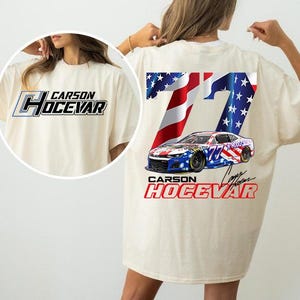 May include: Cream-colored t-shirt featuring a racing car design with the number 77 and the name "Carson Hocevar." The back of the shirt displays an American flag design. The front of the shirt has the name "Carson Hocevar" in a stylized font.