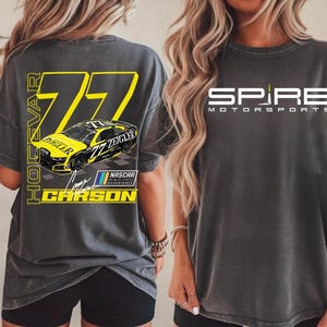 May include: Dark gray t-shirt featuring a racing car design with the number 77 and the name "Carson". The front of the shirt displays the "Spire Motorsports" logo in white. The shirt has short sleeves.
