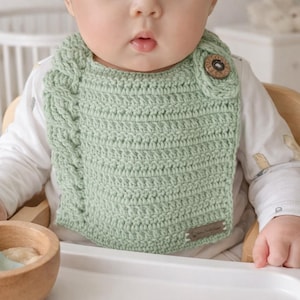 Puede incluir: Babero de bebé de ganchillo verde claro hecho a mano, con un diseño de punto trenzado en el lado izquierdo. El babero tiene un cierre de botón de madera y una pequeña etiqueta marrón. El bebé lleva una camiseta blanca de manga larga.