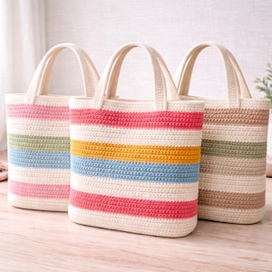 Puede incluir: Tres bolsos de mano de ganchillo con asas y cuerpos color crema. Los bolsos presentan rayas horizontales en varios colores, como rosa, azul, amarillo y verde. Los bolsos están hechos de un material suave y texturizado.