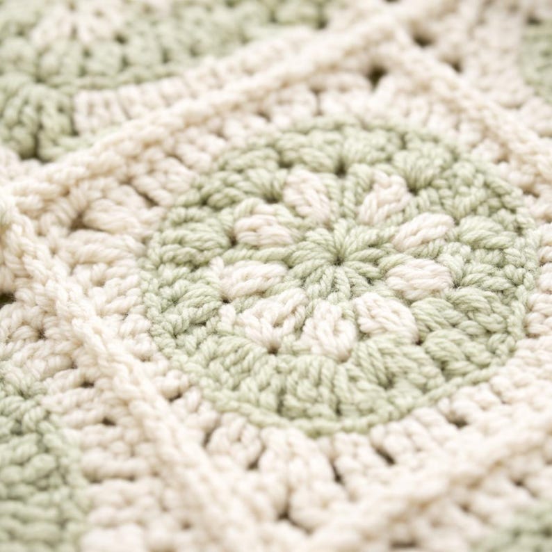 May include: Close-up of a handmade crochet blanket. The blanket features a repeating pattern of squares in soft green and cream colors. The texture of the yarn is visible, creating a cozy and inviting look. The blanket is likely used for home decor.