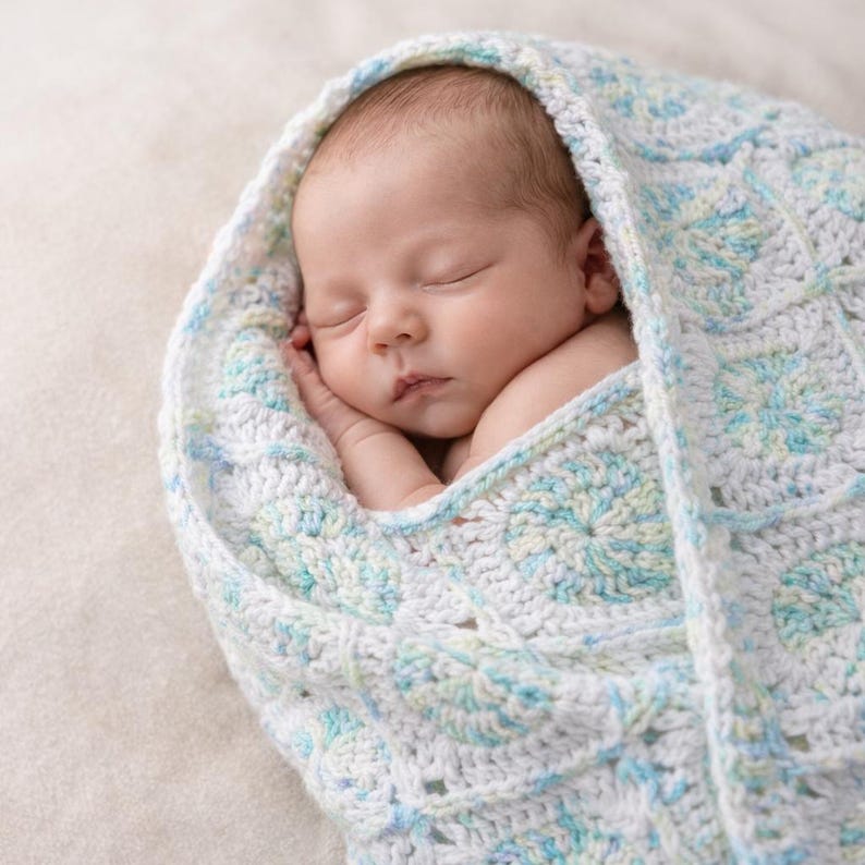 May include: A newborn baby sleeps peacefully, swaddled in a handmade, crocheted blanket. The blanket features a white base with a repeating pattern of blue and light green floral designs. The baby's skin is fair, and the blanket provides a soft, cozy wrap.