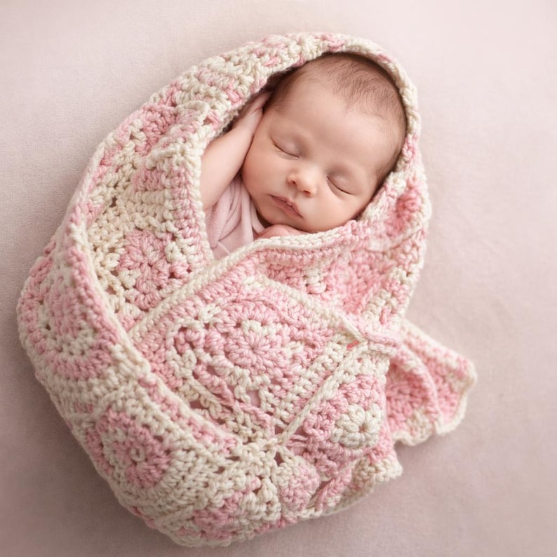 May include: A sleeping baby wrapped in a handmade, pink and cream-colored crochet blanket. The blanket has a floral pattern and a scalloped edge. The baby is wearing a pink onesie.
