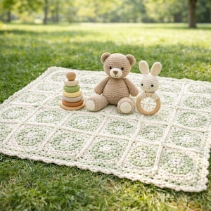 May include: A crocheted baby blanket with a green and white square pattern, a brown teddy bear, a white bunny rattle, and a wooden stacking toy are arranged on a grassy surface. The toys are made of soft materials.