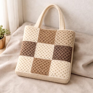 Pode incluir: Bolsa tote de crochê feita à mão com um design de patchwork. A bolsa apresenta um formato quadrado com quadrados creme, marrom claro e marrom escuro. Possui uma alça combinando na cor creme e está sobre uma superfície de cor neutra.