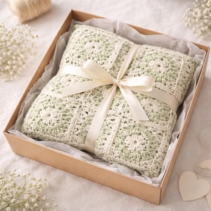 May include: A crocheted blanket in a gift box, tied with a cream-colored ribbon. The blanket features a square pattern in off-white and light green. The box is made of cardboard and lined with tissue paper.