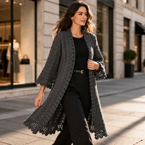May include: A long, open-front gray crochet cardigan with a scalloped hem and sleeves. The cardigan is knee-length and has a delicate, openwork design. The person is wearing a black top and pants with a black belt.