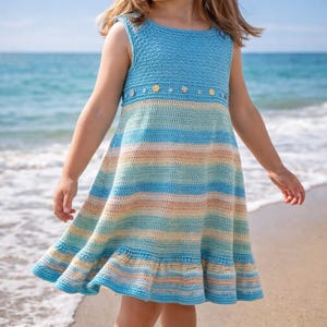 Op de afbeelding: Een mouwloze gehaakte jurk in tinten blauw, bruin en crème. Het lijfje is effen lichtblauw, met kleine bloemaccenten. De rok heeft horizontale strepen en een gefranjerde zoom. De jurk is geschikt voor een kind.