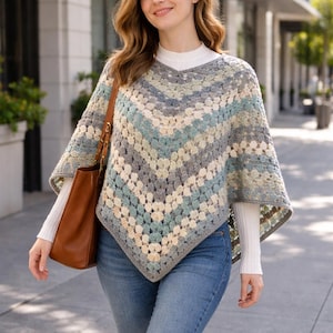 Könnte beinhalten: Ein mehrfarbiger Häkelponcho in Grau-, Blau- und Cremetönen. Der Poncho hat einen V-Ausschnitt und ein geometrisches Muster. Die Person trägt ein weißes Langarmshirt und blaue Jeans. Eine braune Ledertasche wird über der Schulter getragen.