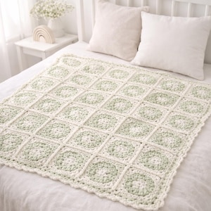 May include: A cream-colored, crocheted blanket with a repeating square pattern. Each square features a central floral design in a soft green hue. The blanket is displayed on a bed with white pillows, suggesting a cozy, handmade home decor item.