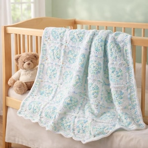 May include: A handmade, white crochet baby blanket with a scalloped edge, featuring a repeating floral pattern in shades of blue and green. The blanket is draped over a wooden crib, next to a teddy bear.