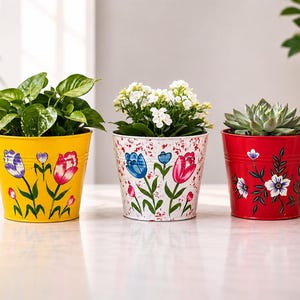 Puede incluir: Tres macetas de metal con diseños florales pintados a mano. La maceta izquierda es amarilla con tulipanes morados y rosas. La central es blanca con motas rojas y tulipanes azules y rosas. La maceta derecha es roja con flores blancas y azules.
