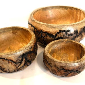 Peut inclure: Trois bols en bois de tailles différentes. Les bols sont en bois brun clair avec des motifs sombres et complexes sur les bords. Les bols sont imbriqués, mettant en valeur leurs différentes tailles et le grain naturel du bois.