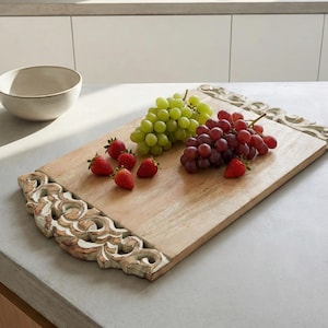 Puede incluir: Tabla de servir rectangular de madera con detalles blancos tallados. Contiene uvas verdes y rojas, y fresas frescas. Un cuenco de cerámica blanca se encuentra en el fondo.