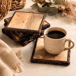 May include: A stack of square wooden coasters with natural bark edges and a light brown wood grain surface. One coaster sits separately in the foreground, supporting a speckled beige ceramic mug filled with dark black coffee.