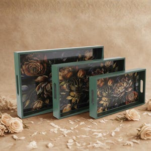 Op de afbeelding: Set van drie rechthoekige serveerbladen. De bladen hebben een donkergroen frame en een bloemenontwerp met rozen en andere bloemen in bruin, geel en blauw. Elk blad heeft uitgesneden handgrepen.