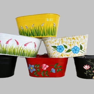 Può includere: Una collezione di sei fioriere ovali in metallo di vari colori, tra cui giallo, bianco, rosso e nero. Ogni fioriera è dipinta a mano con disegni floreali e botanici, come margherite, lavanda e fiori blu. Le fioriere sono impilate.