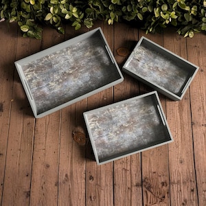 Könnte beinhalten: Set aus drei rechteckigen Serviertabletts mit grauen Rahmen und Innenräumen in Holzoptik. Jedes Tablett hat ausgeschnittene Griffe an den Seiten. Die Tabletts sind auf einer Holzoberfläche angeordnet.