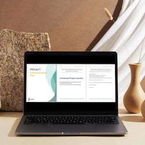 May include: A laptop screen showing a "Project Communication Plan" document with text and a teal and white design. The laptop sits on a beige surface, with vases and a stone block in the background.