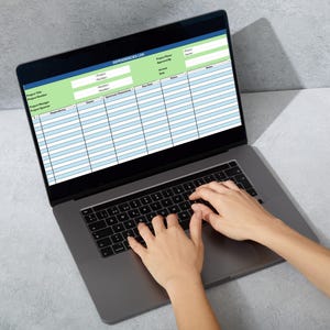 May include: A laptop displaying a spreadsheet with green and blue highlighted sections. The screen shows a table with rows and columns. Hands are typing on the laptop's keyboard. The laptop is on a gray surface.