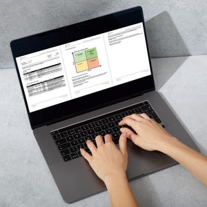 May include: A silver laptop displaying documents with charts and tables. The screen is open, and hands are typing on the keyboard. The laptop is on a gray surface.