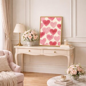 May include: A framed art print featuring a pattern of watercolor hearts in shades of pink and red. The artwork is displayed on a cream-colored background, set in a gold frame. The print is placed on a white console table with a lamp and flowers.