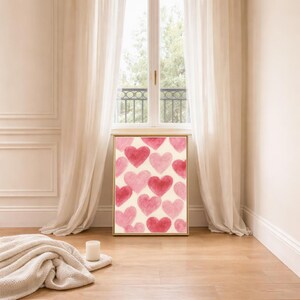 May include: A framed art print featuring a pattern of watercolor hearts in shades of pink and red against a cream background. The artwork is displayed in a room with a window, curtains, and a wooden floor.