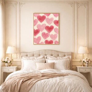 May include: A framed artwork featuring a pattern of watercolor hearts in shades of pink and red against a cream background. The artwork is displayed above a bed with white bedding and a blush-colored throw.