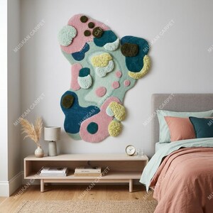 May include: A large, abstract wall hanging rug with a colorful, organic design. The rug features a mix of pink, blue, green, and yellow shapes with textured, raised elements. The rug is mounted on a white wall above a wooden console.