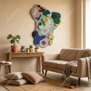 May include: A living room scene featuring a beige sofa, a wooden table with potted plants, and a large, abstract wall hanging in shades of blue, pink, and green. Pillows and a rug complete the cozy, inviting space.