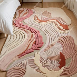 May include: Abstract area rug with a flowing, organic design in shades of pink, beige, and rust. The rug features a textured, raised pattern that creates a three-dimensional effect. The rug is placed on a hardwood floor.