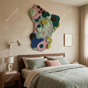 May include: A large, abstract wall hanging rug with a colorful, irregular design. The rug features a mix of textures and colors, including pink, blue, green, and brown. The rug is mounted above a bed with green bedding and a wooden headboard. The brand name "MUSKAN CARPET" is printed on the image.