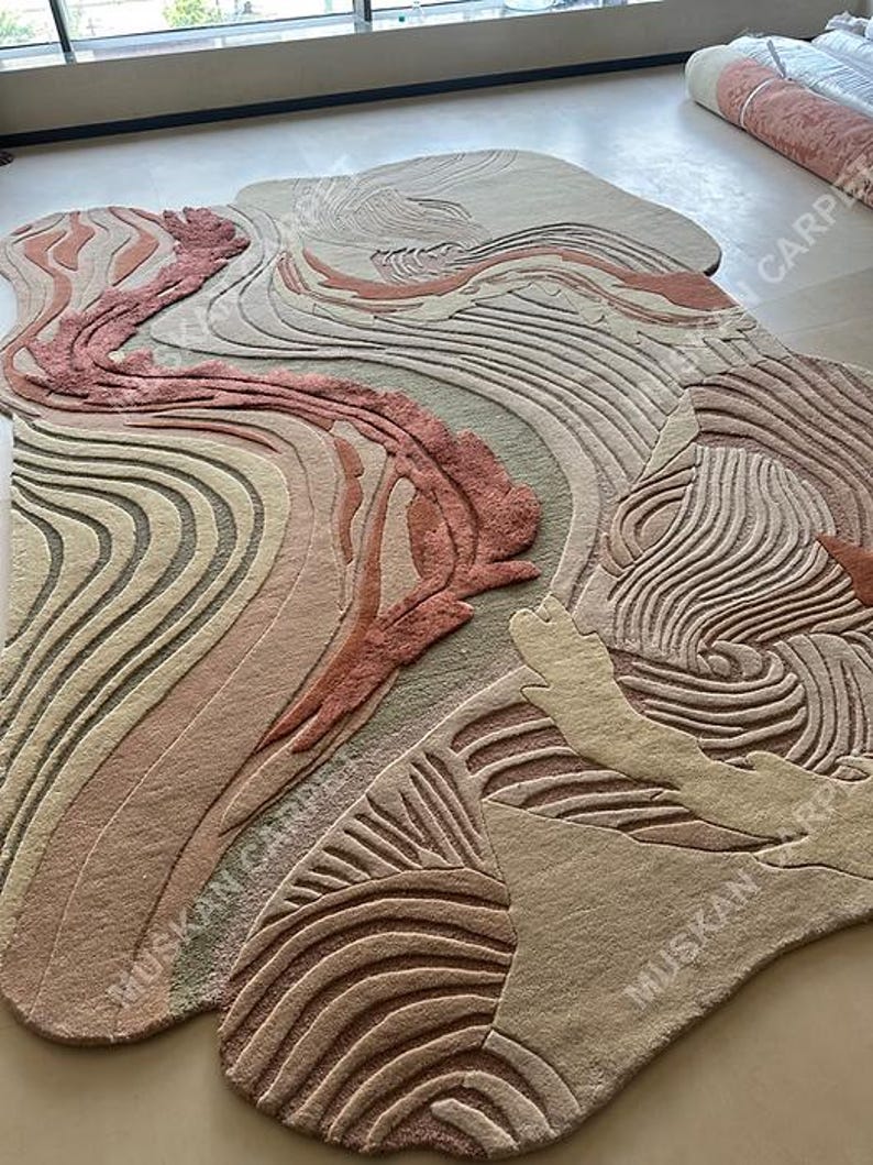 May include: A large, abstract area rug with a flowing, organic design. The rug features a mix of colors, including shades of beige, pink, and brown, with a textured, carved appearance. The design resembles a landscape or geological formation.