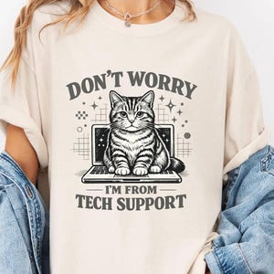 May include: Cream-colored t-shirt featuring a graphic of a cat sitting on a laptop screen. The text above reads "DON'T WORRY", and below, "I'M FROM TECH SUPPORT". The cat is detailed in black and white, with a grid and star design.