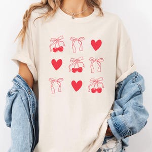 May include: Cream-colored t-shirt featuring a repeating pattern of red cherries, bows, and hearts. The design is printed in a minimalist style, creating a clean and modern aesthetic. The shirt has short sleeves and a relaxed fit.