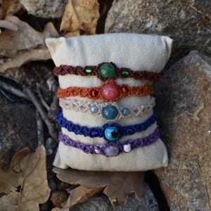 Può includere: Una collezione di braccialetti in macramè fatti a mano in vari colori, tra cui marrone, arancione, blu e viola. Ogni braccialetto presenta una gemma o una perla centrale, con accenti di perline più piccole. I braccialetti sono esposti su un piccolo cuscino bianco sporco.