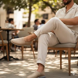 Puede incluir: Zapatos sin cordones de cuero color beige con un diseño perforado en la parte superior. Los zapatos tienen una suela a rayas blanca y azul marino. La persona lleva pantalones beige y una camisa beige, sentada en una silla de madera.