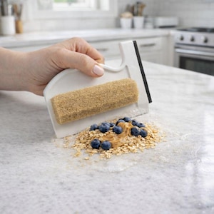 May include: A white kitchen scraper with a handle and a tan scrubbing pad. The scraper is cleaning up a spill of oatmeal, blueberries, and peanut butter on a white countertop. The scraper has a black blade.