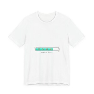 Puede incluir: Camiseta blanca con un gráfico de barra de carga en turquesa, naranja y verde, con el texto "Loading Life...". La barra de carga presenta iconos de una gema, una planta, un libro y un mando de videojuegos.