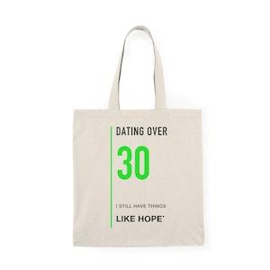 Puede incluir: Una bolsa de tela beige con dos asas. La parte frontal presenta una línea vertical verde junto al texto negro "DATING OVER", un número "30" grande en color verde y el texto negro "I STILL HAVE THINGS LIKE HOPE". El diseño está centrado.