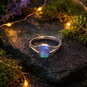 Peut inclure: Une bague en argent ornée d'une pierre d'opale rectangulaire irisée, affichant des nuances de bleu, de violet et de vert. La bague est posée sur une surface sombre et texturée, avec un éclairage doux et de la verdure en arrière-plan. La bague est marquée "925".