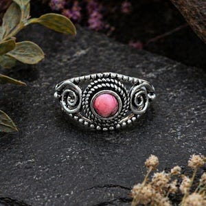 Könnte beinhalten: Ein silberfarbener Ring mit einem rosa Edelstein in der Mitte. Der Ring hat ein detailliertes Design mit Spiral- und Perlenakzenten. Der Edelstein ist rund und hellrosa. Der Ring liegt auf einer dunklen, strukturierten Oberfläche.