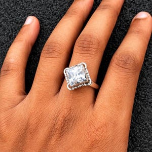 Könnte beinhalten: Ein silberfarbener Ring mit einem großen, quadratisch geschliffenen, klaren Edelstein in einer dekorativen Fassung. Die Fassung verfügt über einen perlenartigen Metallrand um den zentralen Stein, der auf einer glatten, polierten Ringschiene sitzt.