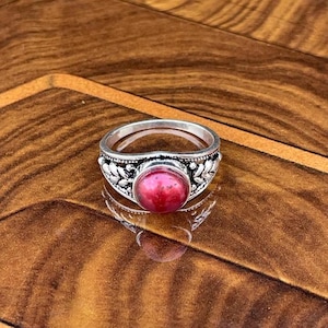 Könnte beinhalten: Ein silberner Ring mit einem großen, runden, roten Edelstein in der Mitte. Der Ring hat ein detailliertes Blumendesign um den Edelstein und entlang des Bandes. Der Ring wird auf einer Holzoberfläche präsentiert.