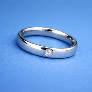 Może przedstawiać: Polerowany srebrny pierścionek z pojedynczym, małym, okrągłym diamentem osadzonym w oprawie. Pierścionek jest prezentowany na jednolitym niebieskim tle, podkreślając jego odblaskową powierzchnię i blask diamentu.