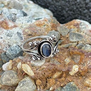 Könnte beinhalten: Ein silberner Ring mit einem ovalen, dunkelblauen Edelstein. Der Ring hat ein verziertes Design mit Wirbelmustern und kleinen, runden Verzierungen. Der Ring ist vor einem strukturierten, felsigen Hintergrund platziert.