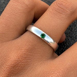 Könnte beinhalten: Ein polierter Silberring mit einem kleinen, runden, smaragdgrünen Edelstein. Der Ring hat ein glattes Band mit einer leichten Krümmung. Der Edelstein ist in der Mitte gefasst und bietet eine minimalistische Ästhetik.