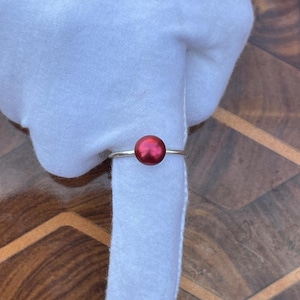 Könnte beinhalten: Ein silberner Serviettenring mit einer großen, runden, roten Perle. Der Ring umschließt eine weiße Stoffserviette und sorgt für eine schlichte, elegante Präsentation. Die Perle hat ein glänzendes Finish.