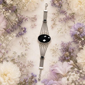 Könnte beinhalten: Ein silberfarbenes mehrreihiges Armband mit einem großen, zentralen, ovalen schwarzen facettierten Stein. Das Design umfasst zwei kleinere schwarze ovale Akzente entlang der Kettenglieder, verbunden durch fünf feine Silberketten und einen Karabinerverschluss.